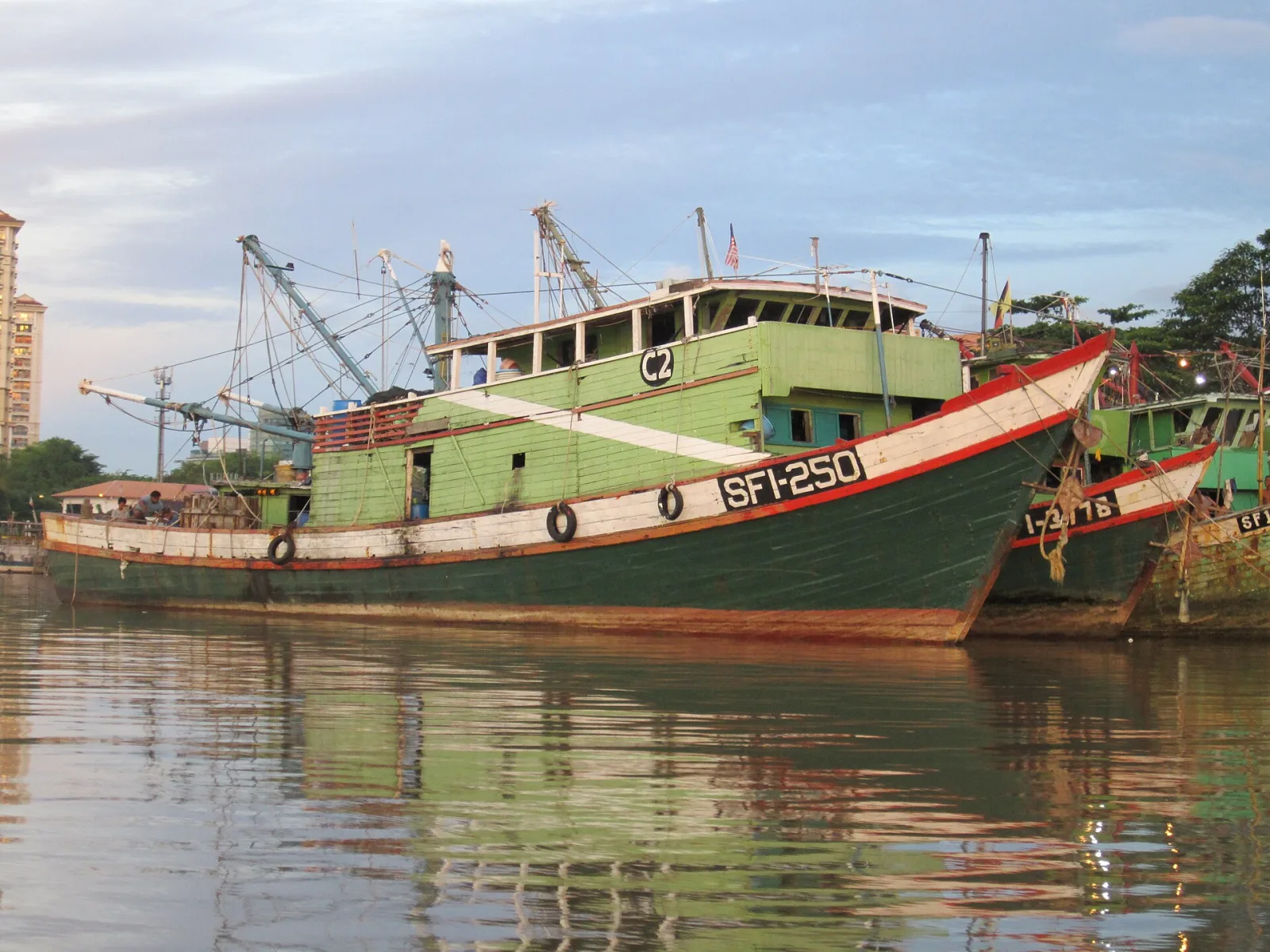 A fishing boat crossing calm Malaysian river water near sunset.